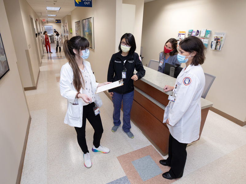 Four healthcare workers are present in a clinic; three stand and talk at the reception desk, and one is positioned behind the desk. Staff and patients are seen in the hallway.